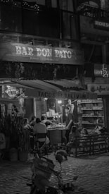 A nighttime street scene with patrons sitting outside a small bar and taco stand. The setting features string lights and a rustic wooden sign with the name 'Bar Don Pato'. Customers are seated around tables enjoying their meals, while a person stands by a grill preparing food. The background shelves hold various products, suggesting a casual dining atmosphere.