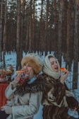 Two people dressed in warm, traditional clothing enjoy food outdoors in a snowy forest setting. A small table in the background holds a samovar and a stack of pastries.