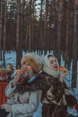 Two people dressed in warm, traditional clothing enjoy food outdoors in a snowy forest setting. A small table in the background holds a samovar and a stack of pastries.