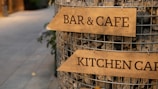 Wooden signs with the words 'BAR & CAFE' and 'KITCHEN CAFE' are attached to a wire structure filled with stones. The setting appears to be an outdoor pedestrian area with a pathway in the background.