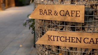 Wooden signs with the words 'BAR & CAFE' and 'KITCHEN CAFE' are attached to a wire structure filled with stones. The setting appears to be an outdoor pedestrian area with a pathway in the background.