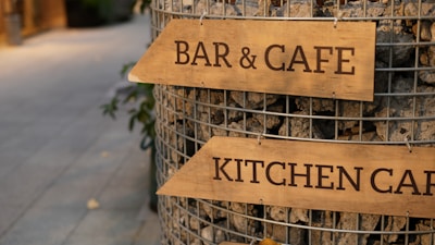 Wooden signs with the words 'BAR & CAFE' and 'KITCHEN CAFE' are attached to a wire structure filled with stones. The setting appears to be an outdoor pedestrian area with a pathway in the background.