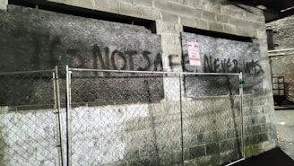 A weathered brick wall with two large dark plywood boards is partially covered with graffiti that reads 'NOT SAFE NEVER WAS.' A metal chain-link fence with a locked gate runs along the wall. A sign on the wall reads 'AUTHORIZED PARKING ONLY.' The area appears neglected, with uneven pavement and dim lighting.