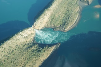 Aerial view of a natural water channel cutting through two land masses. The water appears to be in motion, creating swirling patterns. The surrounding land is covered with sparse vegetation and greenery.