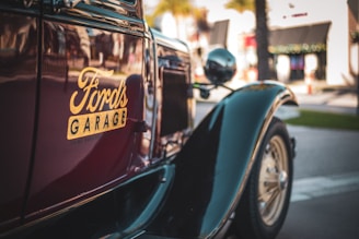 A vintage car with the logo 'Ford's Garage' prominently displayed on its side panel. The car is parked on a street, with blurred buildings and palm trees in the background, indicating an urban setting.