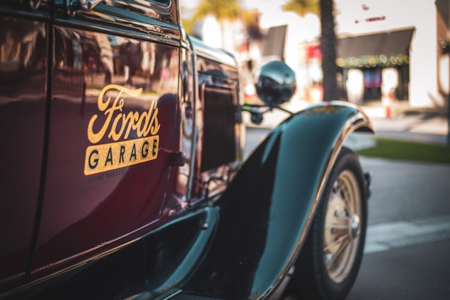 A vintage car with the logo 'Ford's Garage' prominently displayed on its side panel. The car is parked on a street, with blurred buildings and palm trees in the background, indicating an urban setting.