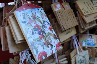 An illustration featuring animated characters with various expressions, surrounded by colorful attire and floral headpieces, is prominently displayed on a wooden plaque. This plaque is among several hanging wooden tablets, each featuring Japanese writing and some vivid illustrations.