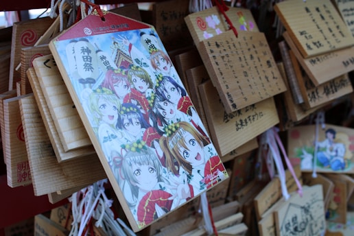 An illustration featuring animated characters with various expressions, surrounded by colorful attire and floral headpieces, is prominently displayed on a wooden plaque. This plaque is among several hanging wooden tablets, each featuring Japanese writing and some vivid illustrations.