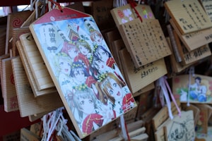An illustration featuring animated characters with various expressions, surrounded by colorful attire and floral headpieces, is prominently displayed on a wooden plaque. This plaque is among several hanging wooden tablets, each featuring Japanese writing and some vivid illustrations.