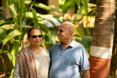 An older couple stands together outdoors, surrounded by lush green foliage, with sunlight filtering through the leaves. The woman is wearing sunglasses and a patterned shawl over her outfit, while the man is adorned in a light blue polo shirt. Both appear to be looking at each other with smiles, creating a warm and affectionate atmosphere.