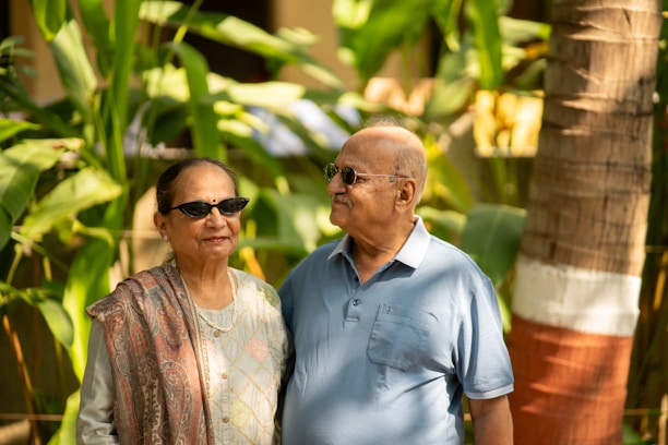 An older LGBTQ couple smiling warmly while sharing a quiet moment in a sunlit garden.
