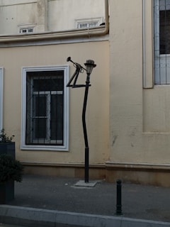 A creatively designed street lamp shaped like a saxophone stands against a beige building wall. The lamp's pole takes the form of a musical instrument, with the light fixture resembling the bell of the saxophone. Nearby, there are a couple of windows with decorative grilles and a small potted plant is partially visible. The ground is paved, and another small post is in the foreground.