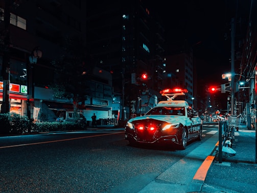 A nighttime urban scene featuring a police car with activated lights parked on a city street. The environment is dimly lit, with neon signs from nearby buildings casting colorful reflections on the pavement. Streetlights and traffic signals are visible, with some pedestrians in the distance and bicycles parked on the sidewalk.
