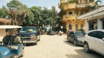 A street scene featuring parked vehicles, including cars and a golf cart, with a yellow building on the right with balconies and a sign. Trees with green foliage line the street, and a few people are walking in the distance. The road surface is cobblestone, and the sky is clear.
