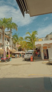 A lively street scene with palm trees lining both sides, various shops with thatched roofs, and a few people walking along the street. A white pickup truck is parked in front of the shops, and traffic cones are placed nearby. The sky is partly cloudy, indicating a warm and tropical atmosphere.
