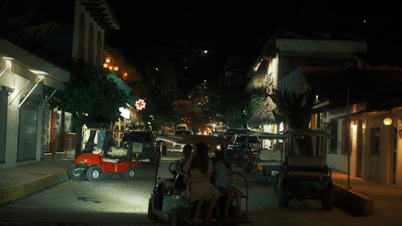 A nighttime street scene with dim lighting and several golf carts parked along the road. Illuminated signs are visible, including a red medical cross. Buildings line the street, featuring exposed lights and trees providing partial cover. Two people are seen sitting on one of the golf carts in the foreground.