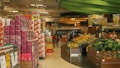 A supermarket aisle filled with stacks of toilet paper and shelves of various products. There are large signs advertising promotions, such as '3x2'. Fresh produce like bananas and watermelons are prominently displayed on the right. The ceiling has green curved structures, and the atmosphere is busy with customers shopping.