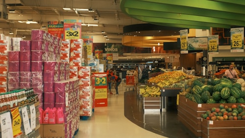 A supermarket aisle filled with stacks of toilet paper and shelves of various products. There are large signs advertising promotions, such as '3x2'. Fresh produce like bananas and watermelons are prominently displayed on the right. The ceiling has green curved structures, and the atmosphere is busy with customers shopping.
