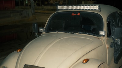 A vintage car with LED light bars mounted on its roof and front. The vehicle appears to be dusty and is parked in a dimly lit outdoor area. The windshield has a decal with the words 'Luna’s C'. The side mirrors and front indicators are visible.