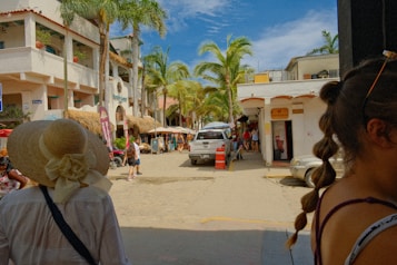 A bustling street scene in a tropical location with palm trees lining the road. A white building with balconies and thatched roofs is on the left. Several people are strolling around, including a person in a large sunhat and another with braided hair. A white SUV is parked on the street, and there are shop signs and canopies.