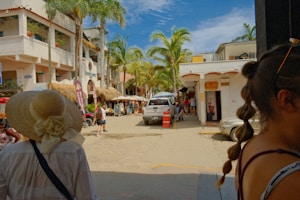 A bustling street scene in a tropical location with palm trees lining the road. A white building with balconies and thatched roofs is on the left. Several people are strolling around, including a person in a large sunhat and another with braided hair. A white SUV is parked on the street, and there are shop signs and canopies.