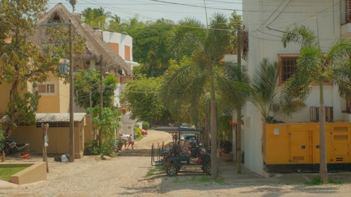 A quiet residential street featuring tropical trees and cobblestone pavement. On the right, there's a bright yellow utility generator, while a black vehicle is parked nearby. The buildings are multi-story structures with white and beige exteriors, and some have thatched roofs. Overhead wires cross the street.