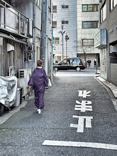 A person wearing a traditional purple kimono walks down a narrow, urban alley lined with buildings. The street is marked with white Japanese characters, and in the background, a black car is parked with several people standing nearby.