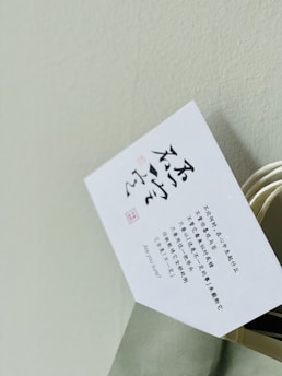A white card with printed text in Chinese characters and some English text, partially inserted into a light green paper bag with a twisted handle. The background is a plain light-colored wall.