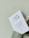 A white card with printed text in Chinese characters and some English text, partially inserted into a light green paper bag with a twisted handle. The background is a plain light-colored wall.