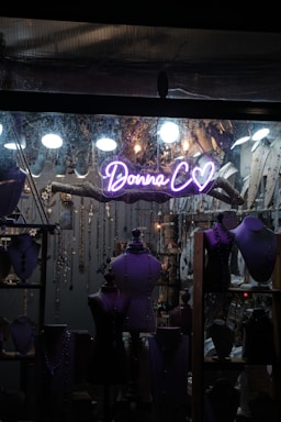 A shop window display featuring several mannequins adorned with jewelry. The backdrop is filled with hanging necklaces and various decorative elements. A neon sign with the words 'Donna C' and a heart symbol is prominently visible in the center.