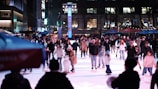 A large group of people are ice skating on a rink in an outdoor urban setting at night. The rink is crowded with skaters of various ages enjoying the activity. In the foreground, a snowman figure can be seen on the ice. Buildings and trees with illuminated lights provide the backdrop, adding to the festive atmosphere.
