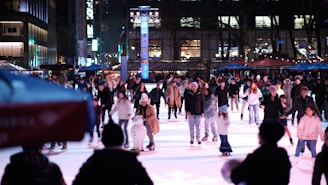 A large group of people are ice skating on a rink in an outdoor urban setting at night. The rink is crowded with skaters of various ages enjoying the activity. In the foreground, a snowman figure can be seen on the ice. Buildings and trees with illuminated lights provide the backdrop, adding to the festive atmosphere.