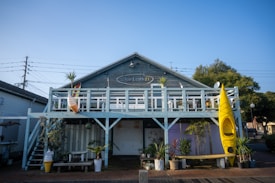 A two-story wooden building with a rustic appearance, featuring a balcony lined with potted plants and outdoor decor. A bright yellow kayak is vertically mounted against the railing, and below, a colorful surfboard adds to the outdoor theme. An oversized ice cream cone sculpture suggests a casual, laid-back setting, and small tables and benches are scattered around.