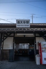 An exterior view of a train station with a sign reading Nishi-Ōita Station. The image captures a rustic building structure with wooden beams and a traditional tiled roof. In the foreground, a vending machine is visible next to a trash can. The sky is clear, and power lines stretch across the view, adding an urban feel.