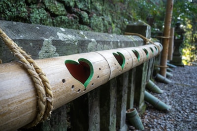 A series of bamboo sections are aligned horizontally, featuring heart-shaped cutouts with a green background. The bamboo is tied together with rope and set against a moss-covered stone wall, creating a natural and rustic scene.
