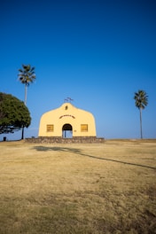 A small, yellow chapel with a cross on top sits on a grassy hill. There are palm trees on either side of the chapel, and the sky is clear blue. The chapel has a stone base and two closed wooden windows on either side of the entrance.
