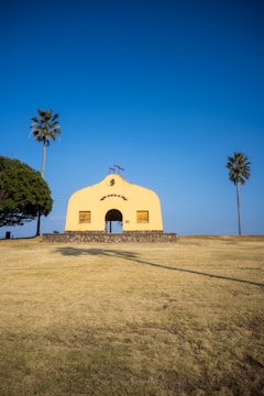 A small, yellow chapel with a cross on top sits on a grassy hill. There are palm trees on either side of the chapel, and the sky is clear blue. The chapel has a stone base and two closed wooden windows on either side of the entrance.