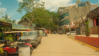 A street scene with parked vehicles including a golf cart and a four-wheeler. Various buildings line the street, some with thatched roofs, and people are walking around. Green trees and banners with decorative elements add color to the scene.