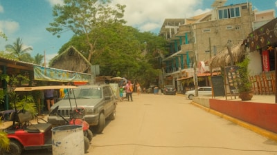 A street scene with parked vehicles including a golf cart and a four-wheeler. Various buildings line the street, some with thatched roofs, and people are walking around. Green trees and banners with decorative elements add color to the scene.