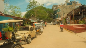 A quaint street scene with several parked vehicles, including a beige vintage car and a golf cart. Small shops and cafes line the street, with a variety of architectural styles. People are casually walking along the sidewalk, and motorcycles are parked nearby. The sky is partly cloudy with lush green trees providing a backdrop.
