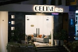 A modern hotel entrance with a well-lit sign reading 'CELINE Hotel'. The exterior features sleek, dark marble walls and glass doors, leading to a cozy lobby area with a gray sofa adorned with orange and gray cushions. Potted plants flank the entrance, adding a touch of greenery.