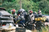 A group of motorcyclists is parked beside a road, each equipped with helmets and riding gear. Brightly colored backpacks and bags are strapped to the backs of the motorcycles. A nearby parked car is visible, partially obscured by foliage and the riders.