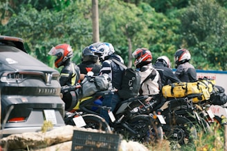 A group of motorcyclists is parked beside a road, each equipped with helmets and riding gear. Brightly colored backpacks and bags are strapped to the backs of the motorcycles. A nearby parked car is visible, partially obscured by foliage and the riders.