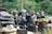 A group of motorcyclists is parked beside a road, each equipped with helmets and riding gear. Brightly colored backpacks and bags are strapped to the backs of the motorcycles. A nearby parked car is visible, partially obscured by foliage and the riders.