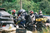 A group of motorcyclists is parked beside a road, each equipped with helmets and riding gear. Brightly colored backpacks and bags are strapped to the backs of the motorcycles. A nearby parked car is visible, partially obscured by foliage and the riders.