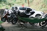 A row of parked motorcycles and scooters are situated on a dirt area, with a white pickup truck in the background. A green hammock is set up between some of the motorcycles, suggesting a casual and outdoor setting. Trees and bushes provide a natural backdrop.