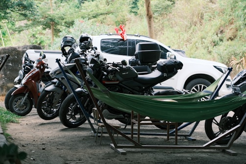 A row of parked motorcycles and scooters are situated on a dirt area, with a white pickup truck in the background. A green hammock is set up between some of the motorcycles, suggesting a casual and outdoor setting. Trees and bushes provide a natural backdrop.