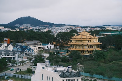 A panoramic view of a community with a large, ornate yellow pagoda surrounded by modern residential houses and greenery. In the background, there is a mountain range under an overcast sky. The houses are mostly a mix of white and blue, and the landscape includes trees and additional buildings.