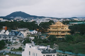 A panoramic view of a community with a large, ornate yellow pagoda surrounded by modern residential houses and greenery. In the background, there is a mountain range under an overcast sky. The houses are mostly a mix of white and blue, and the landscape includes trees and additional buildings.