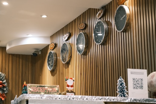 Several clocks with different time zones are mounted on a wooden panel wall. A reception counter is adorned with festive decorations, including a small Santa figurine and miniature Christmas trees. A sign reading 'RECEPTION' is prominently displayed alongside a QR code on a piece of paper.